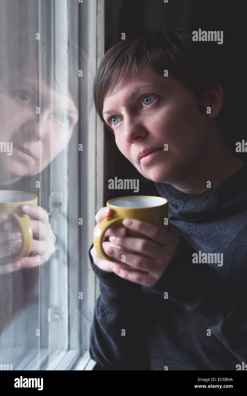 Lonely woman drinking cup of coffee by the window of her living room, looking out with a sad look on her face. Selective focus w Stock Photo