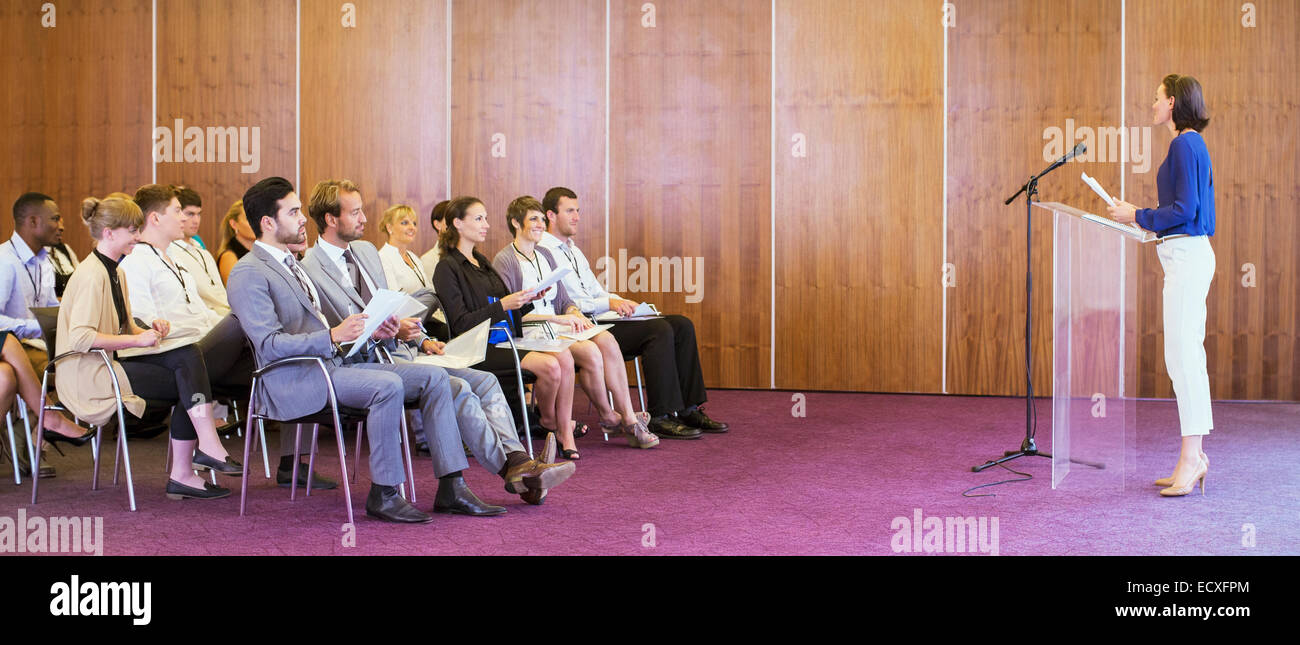 Young woman standing at transparent lectern, talking before audience in conference room Stock Photo