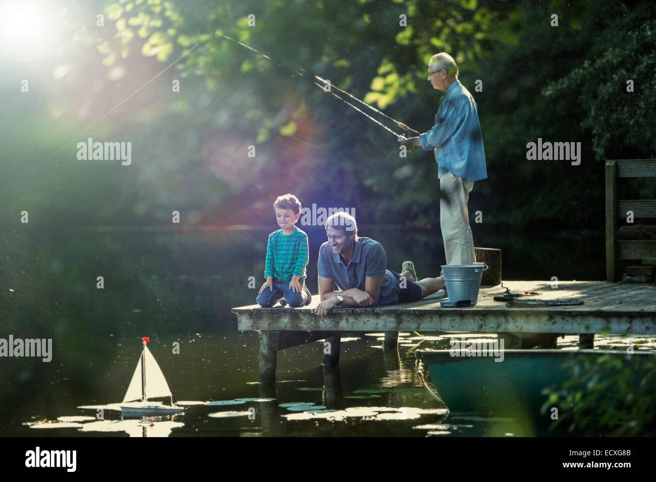 Boy fishing and playing with toy sailboat with father and grandfather at lake Stock Photo