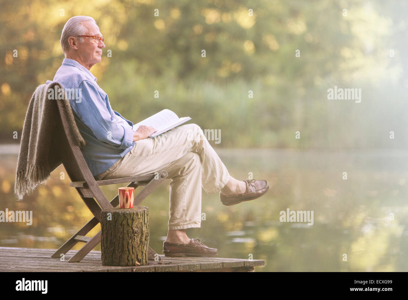 Older man reading book on dock at lake Stock Photo