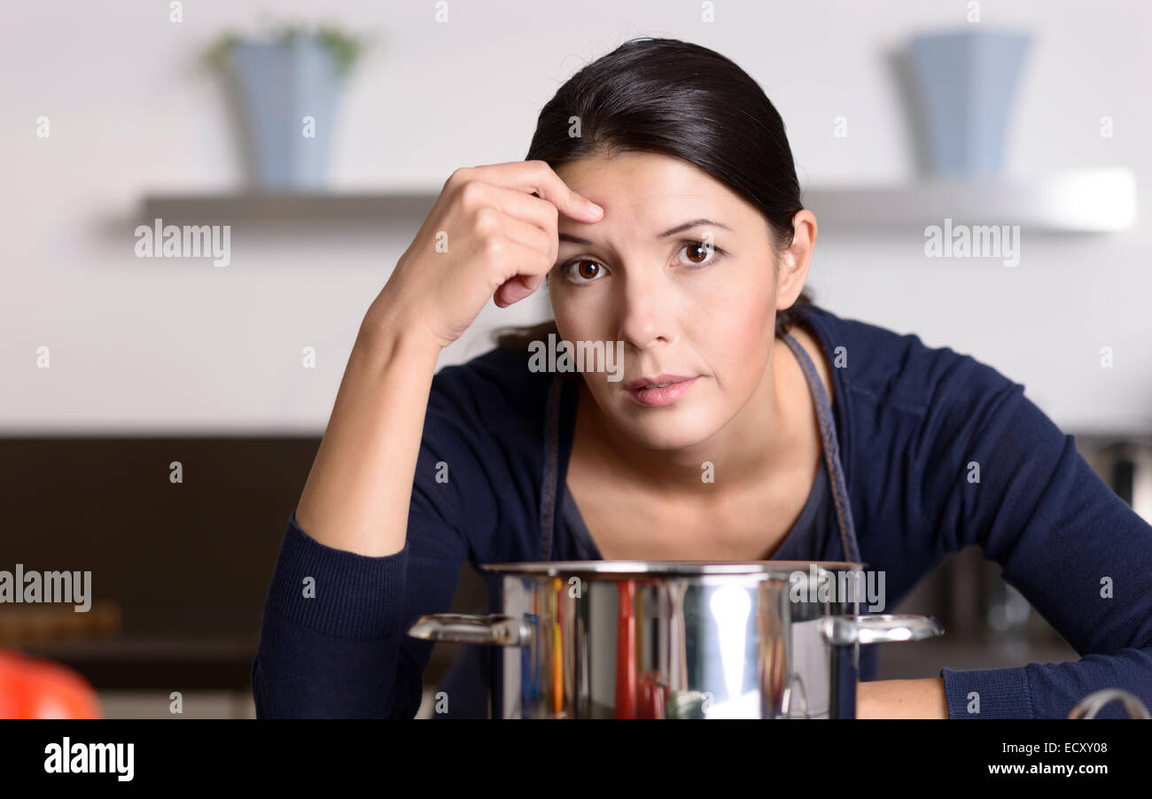 Unmotivated attractive young woman preparing the dinner leaning on the hob eyeing the camera with a listless glum expression as  Stock Photo