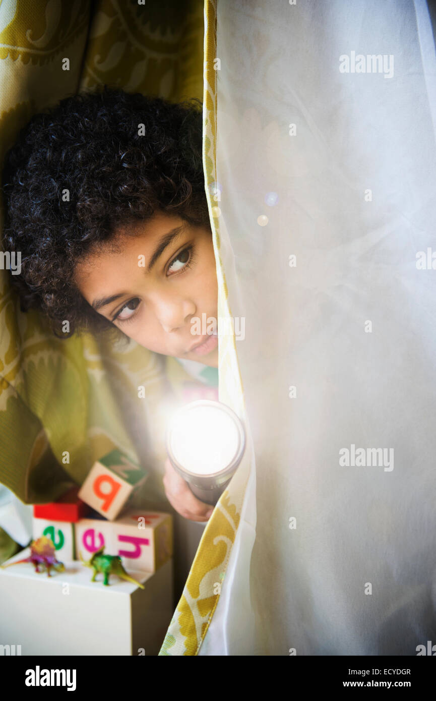 Mixed race boy with flashlight peering out of blanket fort Stock Photo