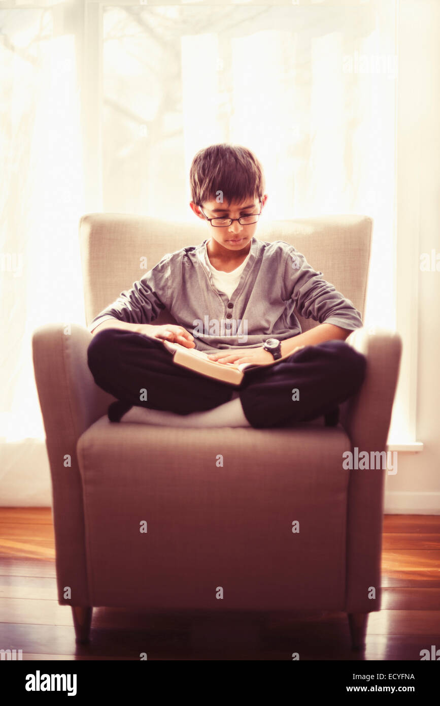 Mixed race boy reading book in armchair Stock Photo