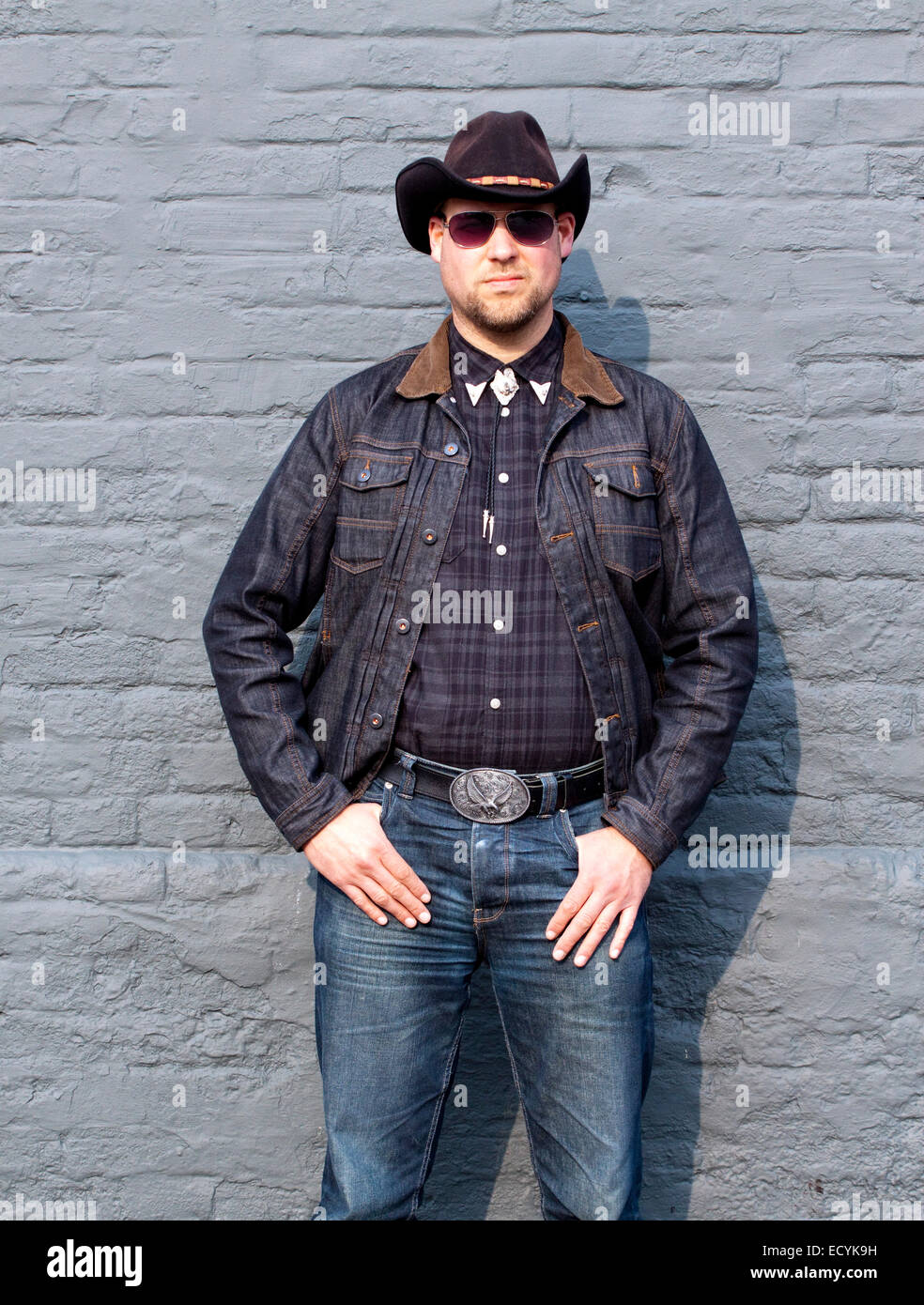 A young man dressed as a cowboy and wearing sunglasses poses in a macho ...