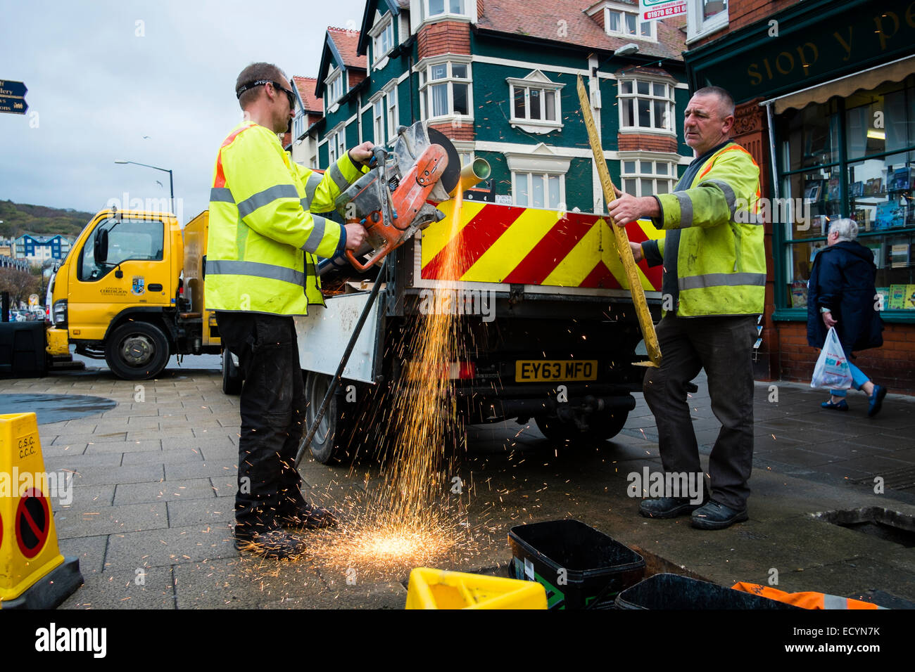 A two-man crew of Ceredigion County Council local authority direct labour workers wearing hi-vis yellow jackets cutting a metal pole with an angle grinder in the street, Aberystwyth Wales UK Stock Photo