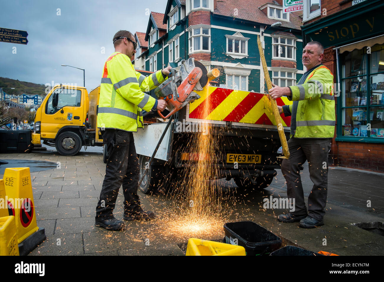 A two-man crew of Ceredigion County Council local authority direct labour workers wearing hi-vis yellow jackets cutting a metal pole using an angle grinder in the street, Aberystwyth Wales UK Stock Photo