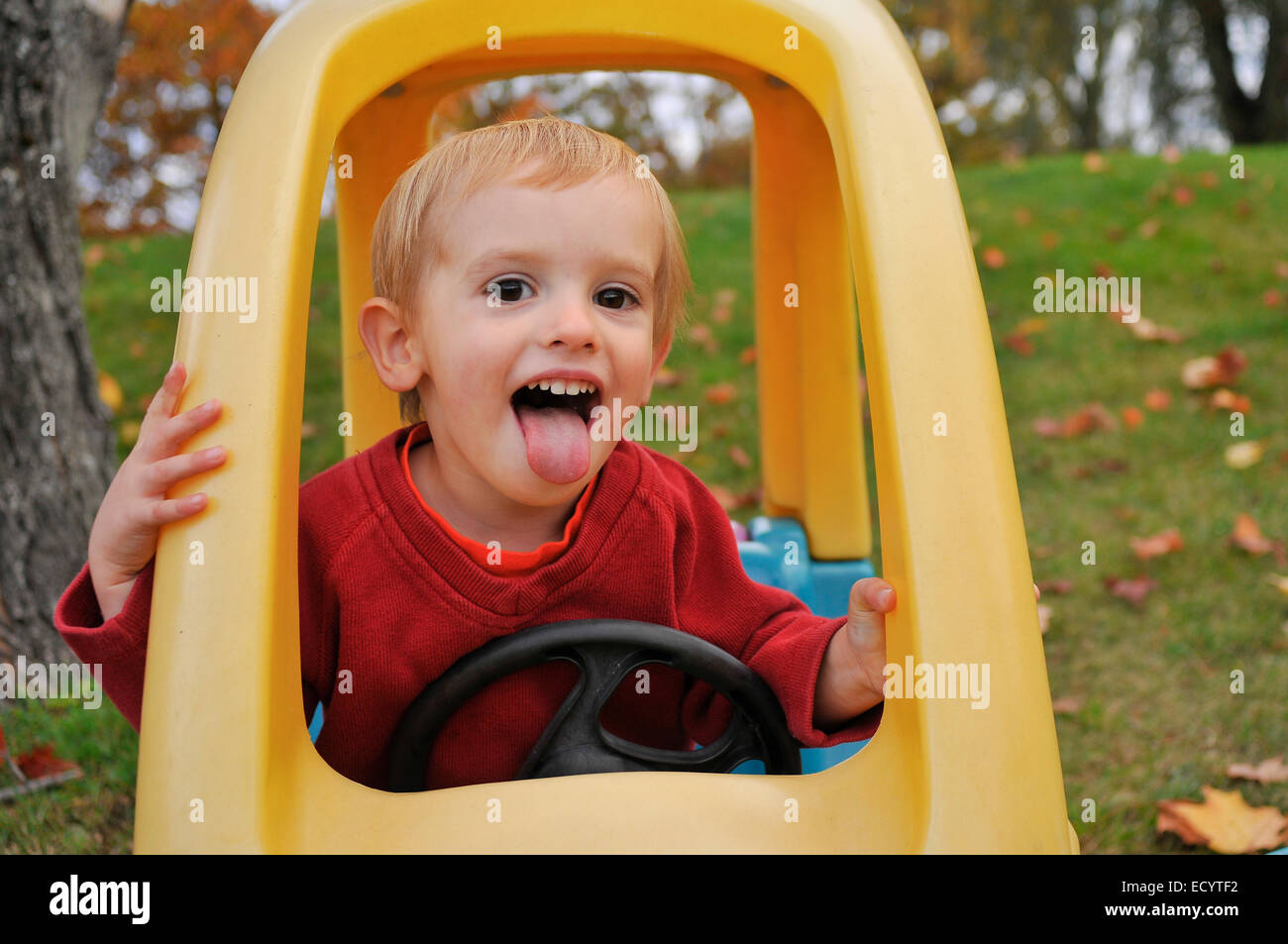 A two-year-old boy makes a funny face while playing in his toy car on an autumn day. Stock Photo