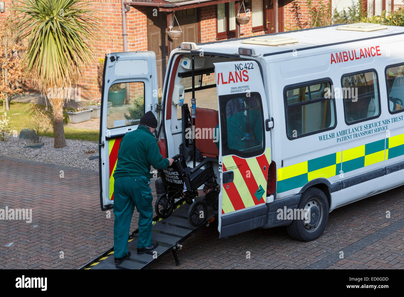 Welsh Ambulance NHS Trust patient transport service man pushing a wheelchair up ramp outside a house after taking a patient home Stock Photo