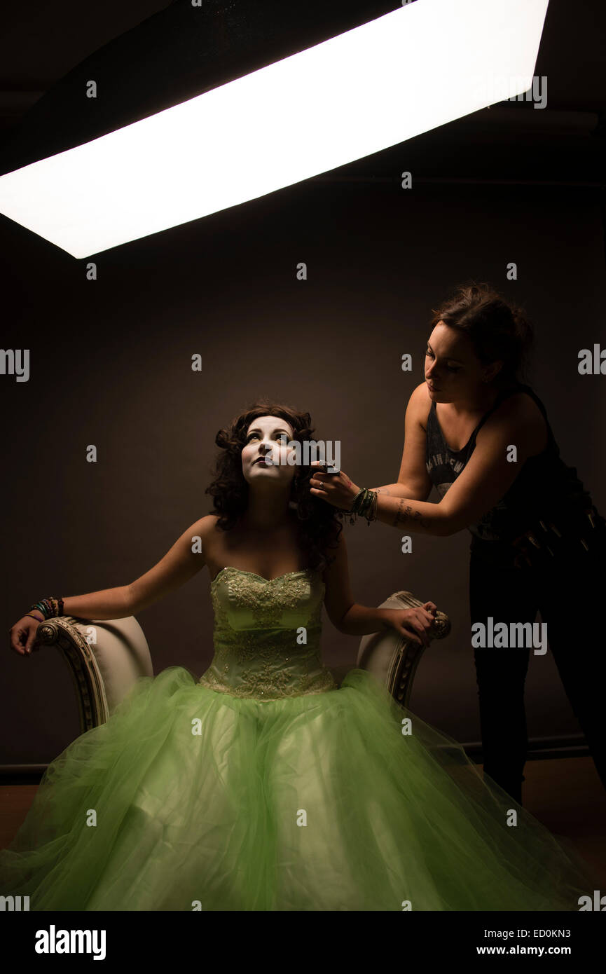 Fantasy makeover photography: A young woman girl model being made up by a make up artist to look like a porcelain white-faced painted puppet and posing like a doll in a photo studio wearing a big green frock dress ball gown Stock Photo