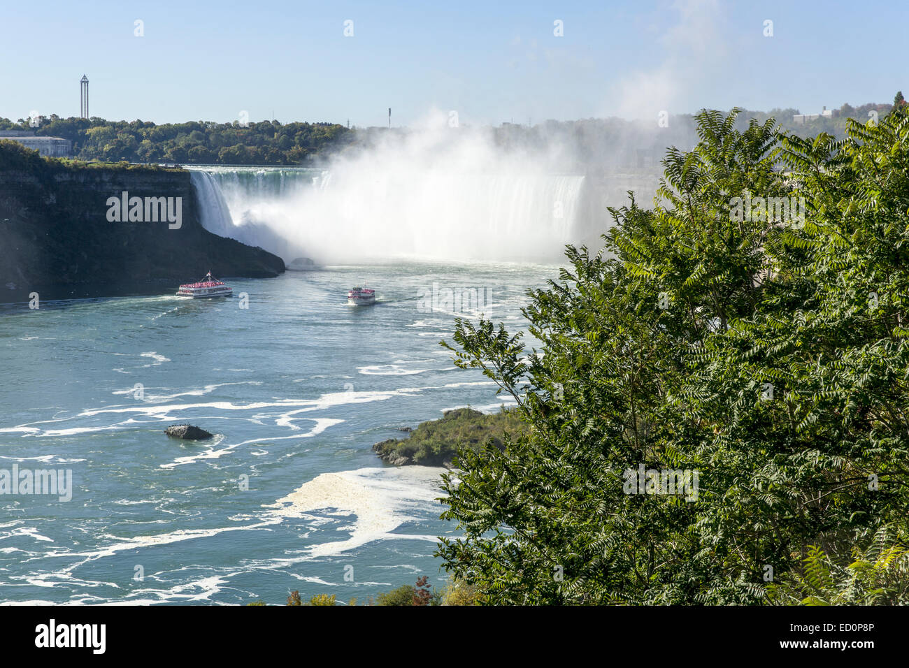Niagra Falls showing the tourist boats with spray, blue sky and foreground foliage. Stock Photo