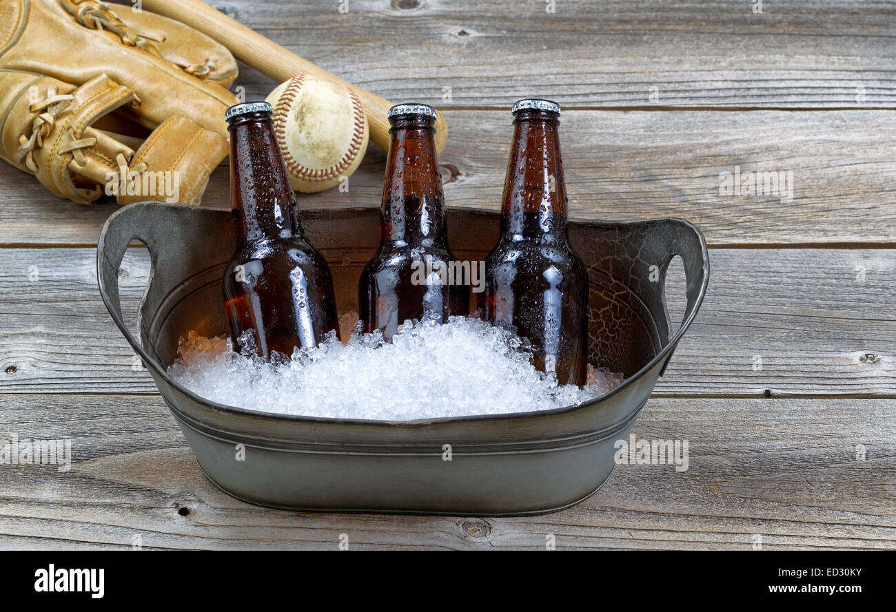 Front view of three brown bottled beers, crushed ice in metal bucket, and baseball equipment in background on rustic wood Stock Photo
