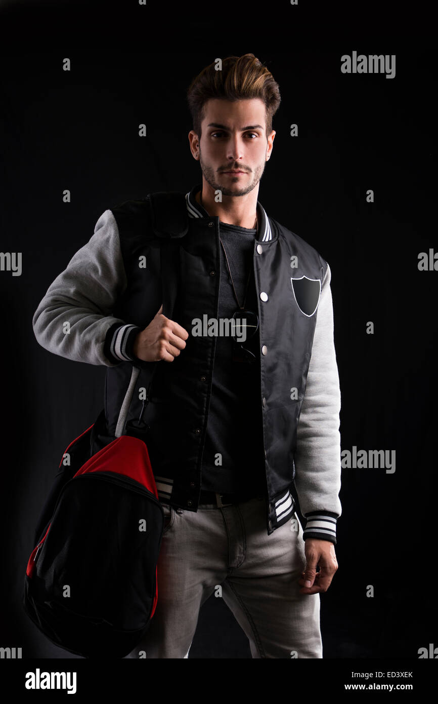 Close up Gorgeous Man in Trendy Fashion Outfit Wearing University Jacket with Sport Bag. Captured in Studio on Black Background. Stock Photo
