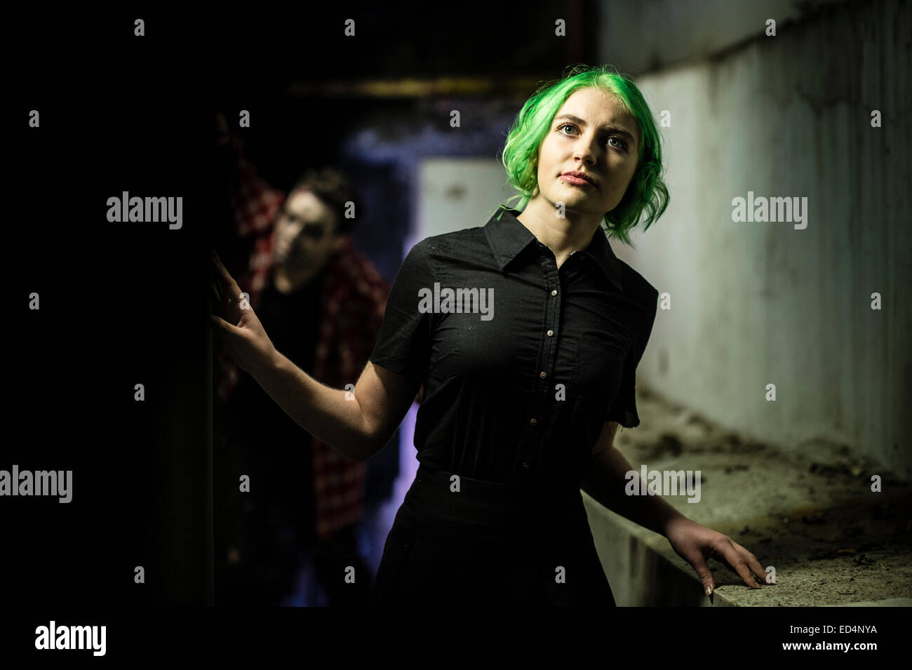 Fantasy photoshoot: makeup and theatre design students role playing a 'murder mystery' stalking movie scene - a young green haired woman girl being stalked followed in a dark alley at night UK Stock Photo