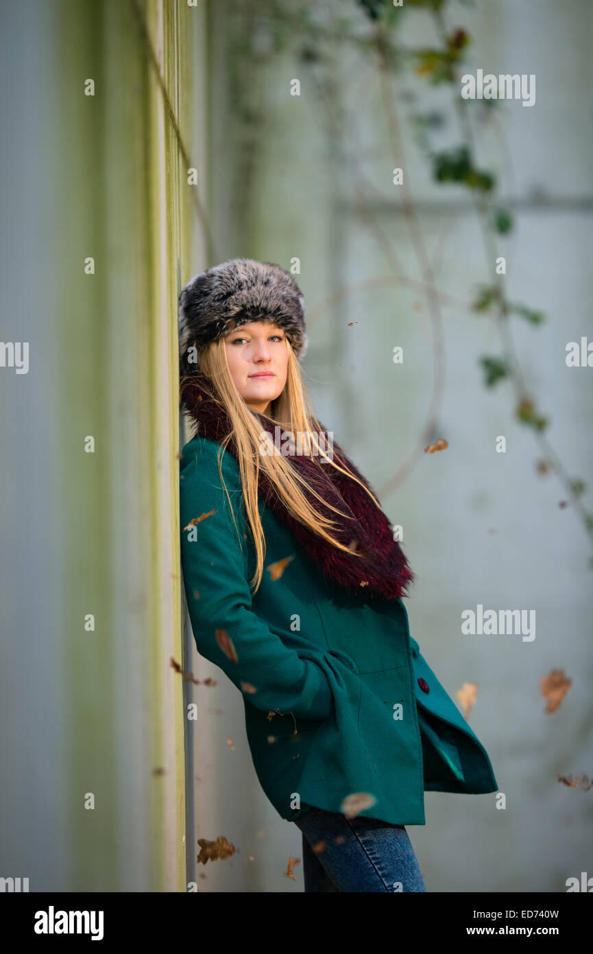 a young cool 'scandinavian' blonde woman teenage girl wearing a green coat and fur hat alone leaning back against a concrete wall outdoors  on a cold winter day UK Stock Photo