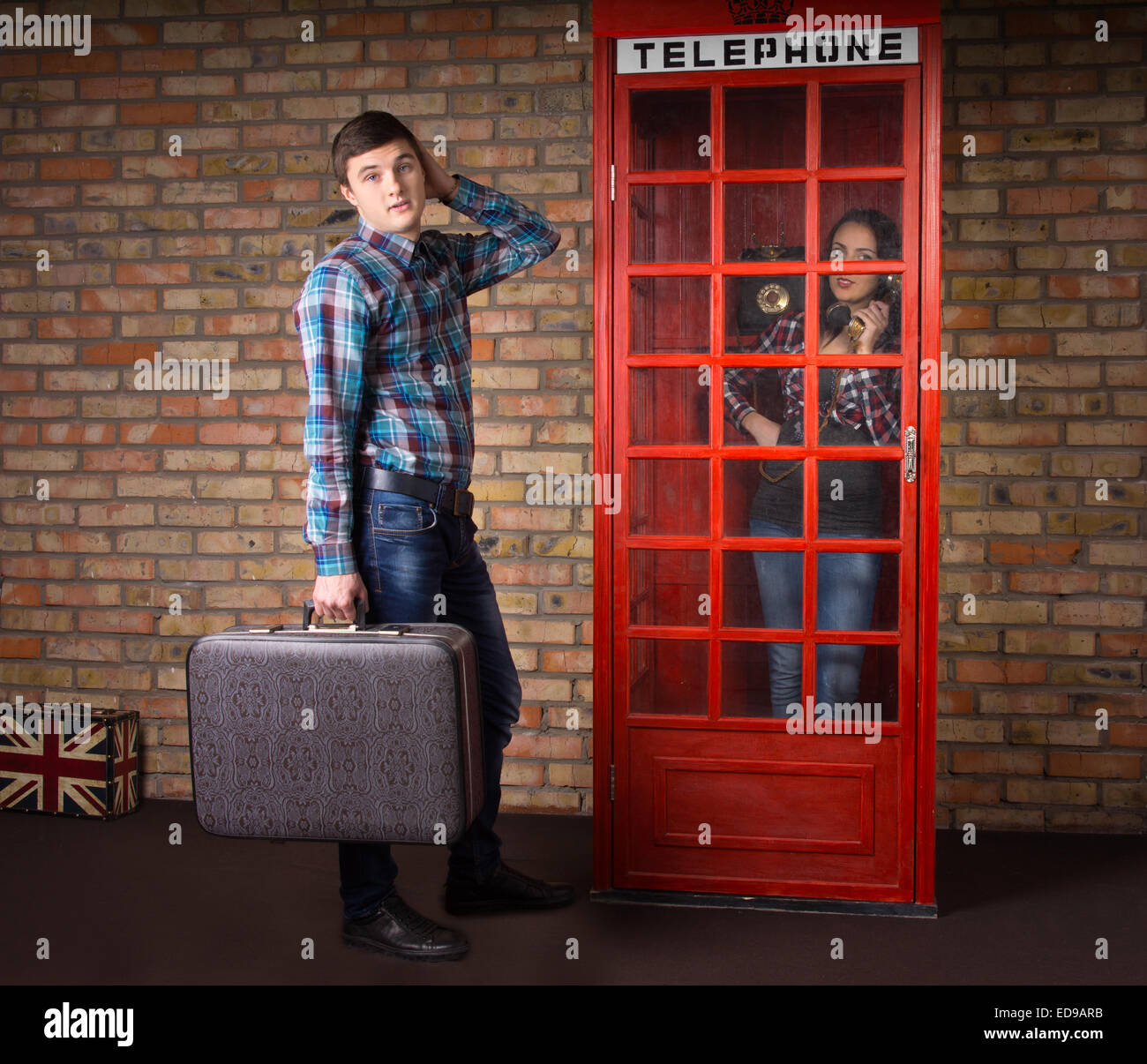 Young Handsome Man Carrying Suitcase Waiting at the Telephone Booth with Woman Using the Telephone Inside. Stock Photo