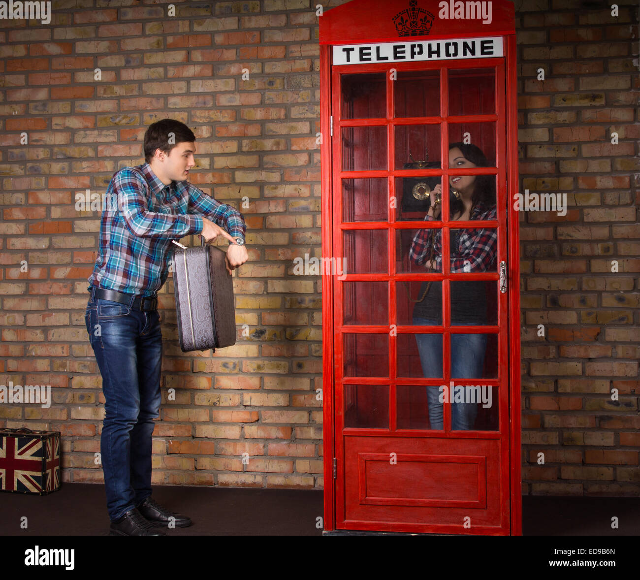 Impatient man pointing to his watch as he stands alongside a red British telephone booth with his suitcase while his wife chats Stock Photo