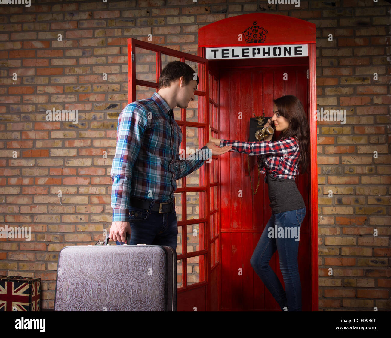 Young man imploring his wife to get off the phone as he stands outside the telephone booth with his suitcase in his hand Stock Photo