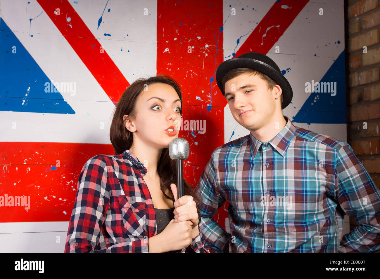 Close up Young Woman with Microphone, Showing Wide Open Eyes Expression, Interviewing Young Male Guest in Trendy Outfit in Front Huge British Flag Print.. Stock Photo