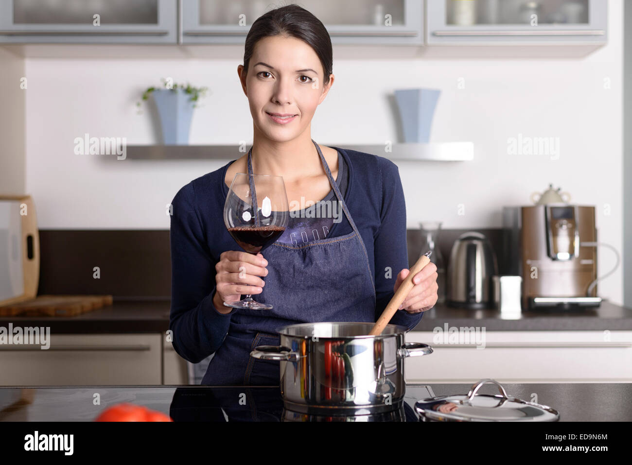 Smiling young housewife celebrating with red wine raising her glass to toast the camera as she stands at the stove Stock Photo