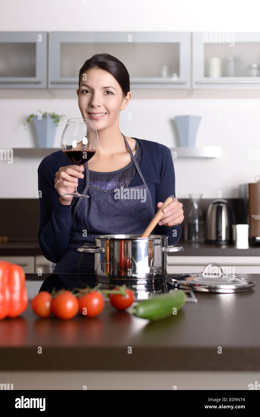 Smiling young housewife celebrating with red wine raising her glass to toast the camera as she stands at the stove Stock Photo