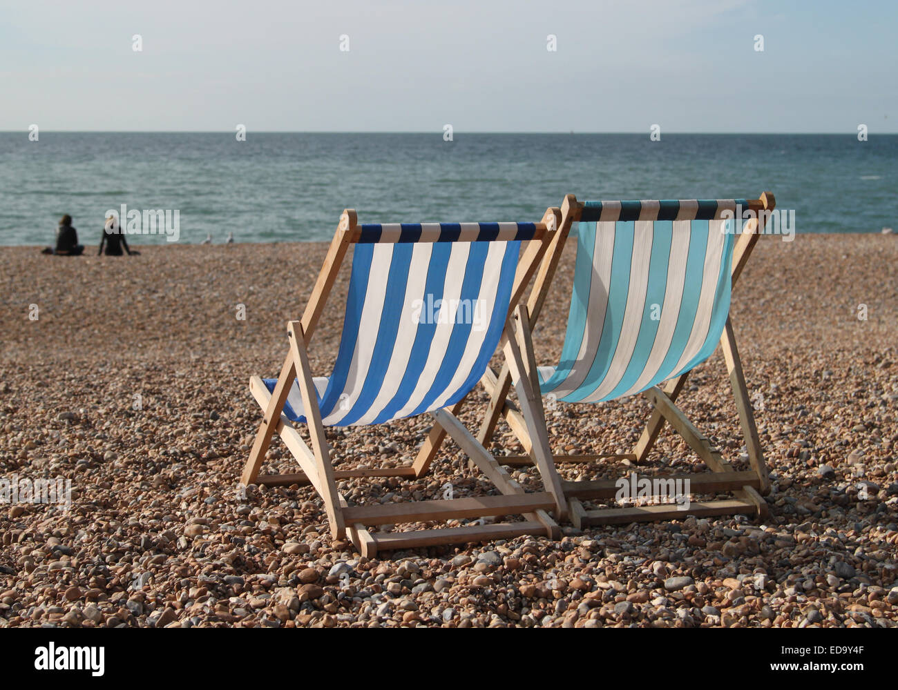 Two empty deck chairs on Brighton beach with two people and sea in the ...