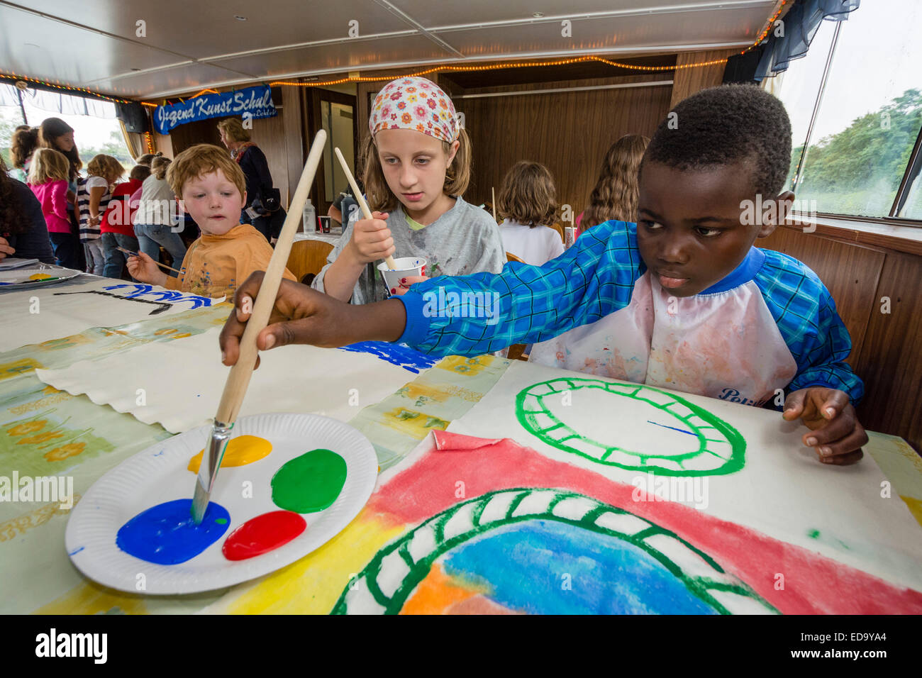 Six to ten year old black and white children enjoy a trip with a school of arts for kids, creating a painting. Stock Photo