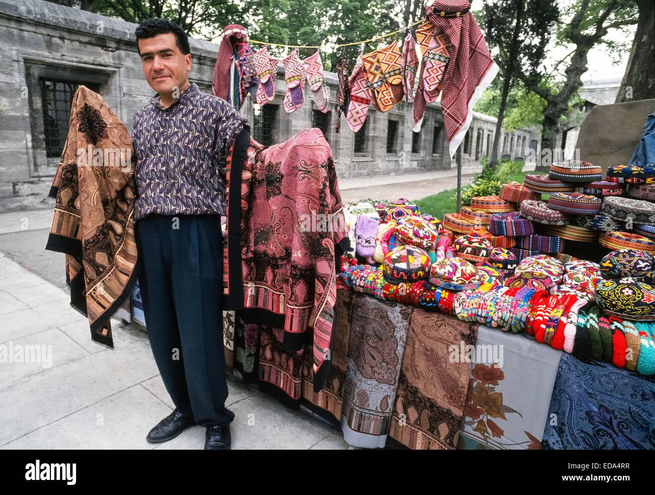 A male vendor displays patterned shawls and very colorful knit caps and
