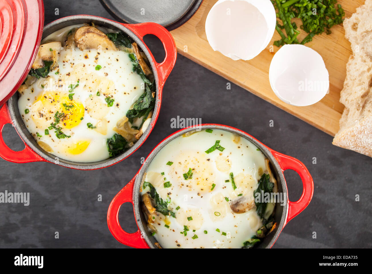 Baked Eggs in red Le Creuset croquettes on slate with garnishes and cutting board background Stock Photo