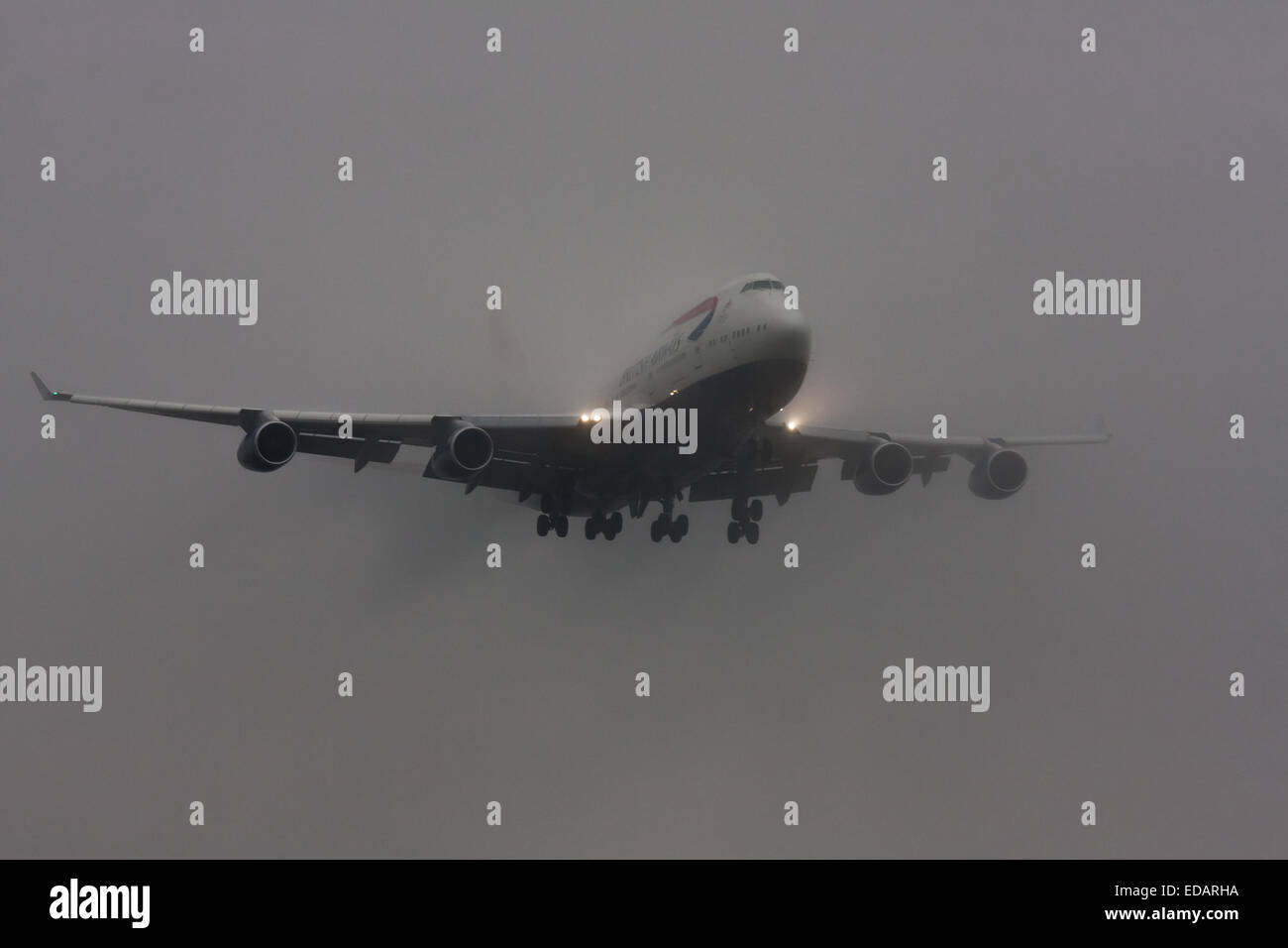 January 3rd 2015, Heathrow Airport, London. Low cloud and rain provide ideal conditions to observe wake vortexes and 'fluffing' as moisture condenses over the wings of landing aircraft. With the runway visible only at the last minute, several planes had to perform a 'go-round', abandoning their first attempts to land. PICTURED: A British Airways Boeing 747 Jumbo Jet emerges from the low cloud moments before touchdown on Heathrow's runway 27L. Stock Photo