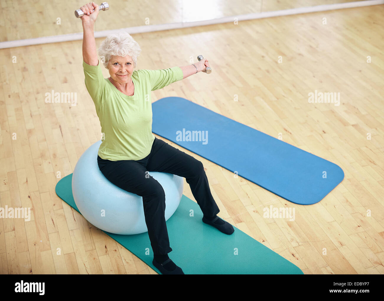Active senior woman sitting on a pilates ball and lifting dumbbells looking at camera. Old Caucasian woman exercising with weigh Stock Photo