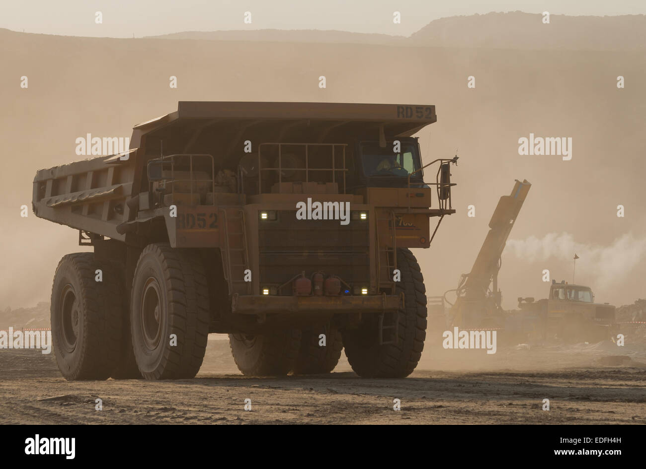 A large Caterpillar mining truck drives across a dusty open cast mine with a drill rig in the background. Stock Photo
