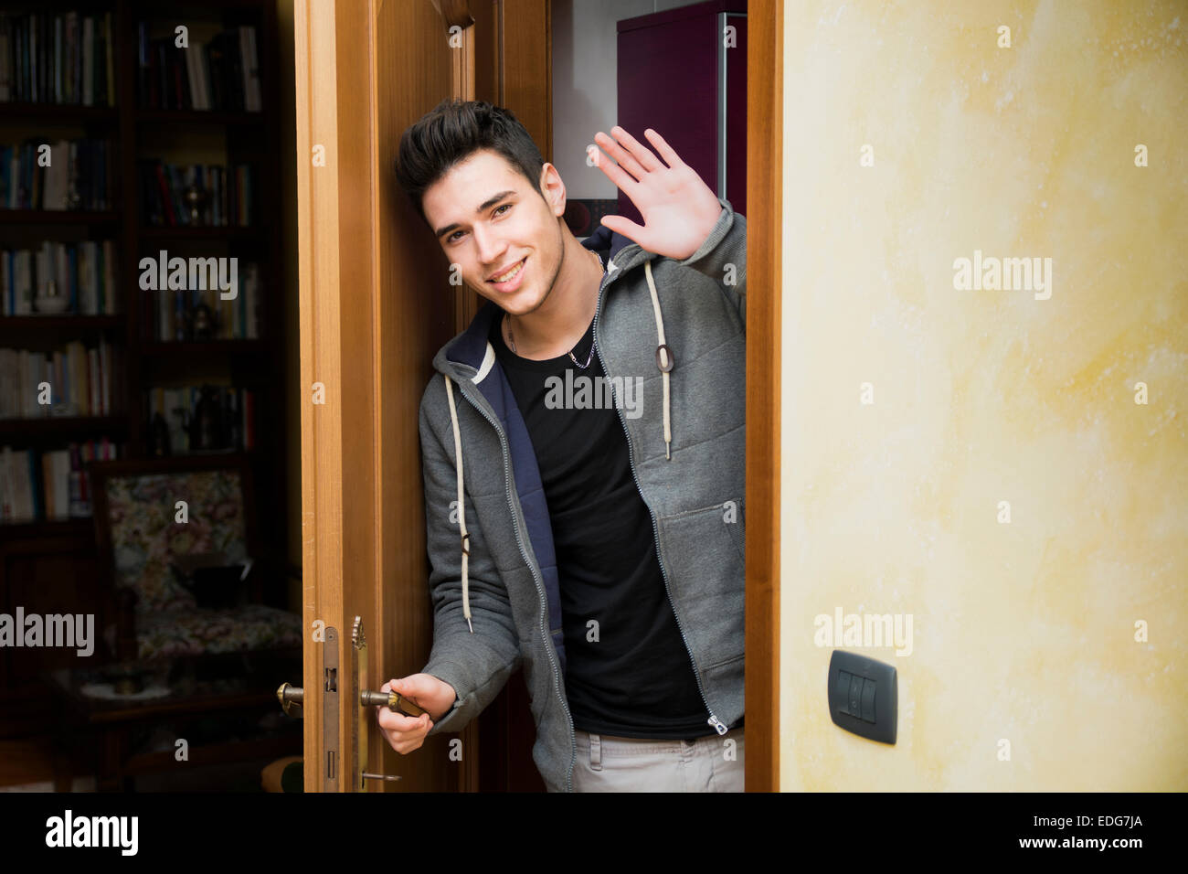 Smiling young man getting out of door waving at the camera with a friendly cheerful smile as he peers around the edge of a woode Stock Photo