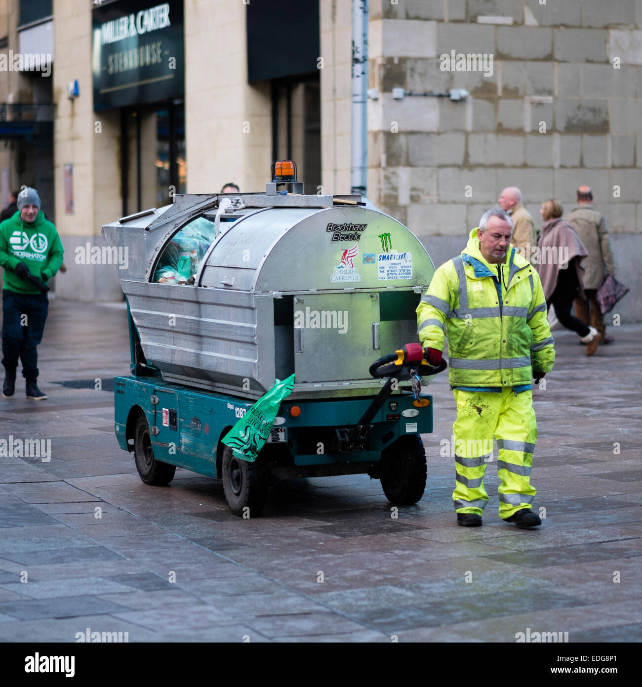 A street cleaner working pulling an electric rubbish cart trolley in