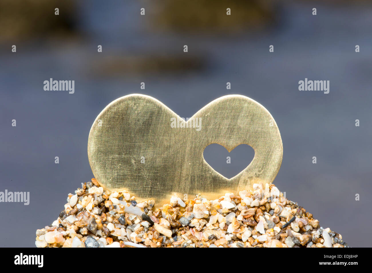 A bright beautiful metal heart is placed on top of a small sand hill. In the background is a rocky beach and waves. Stock Photo