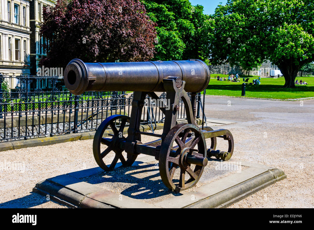 A black cast iron cannon mounted on its carriage stands in the grounds of Rochester castle with green trees and parkland behind Stock Photo