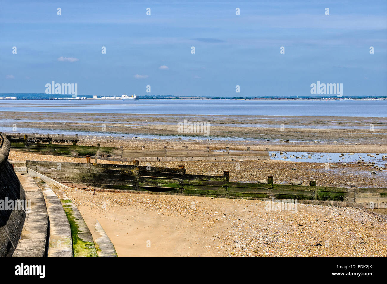A tranquil River Thames flows past a muddy shingle beach as the sun reflects from petrochemical plant on the opposite shoreline Stock Photo