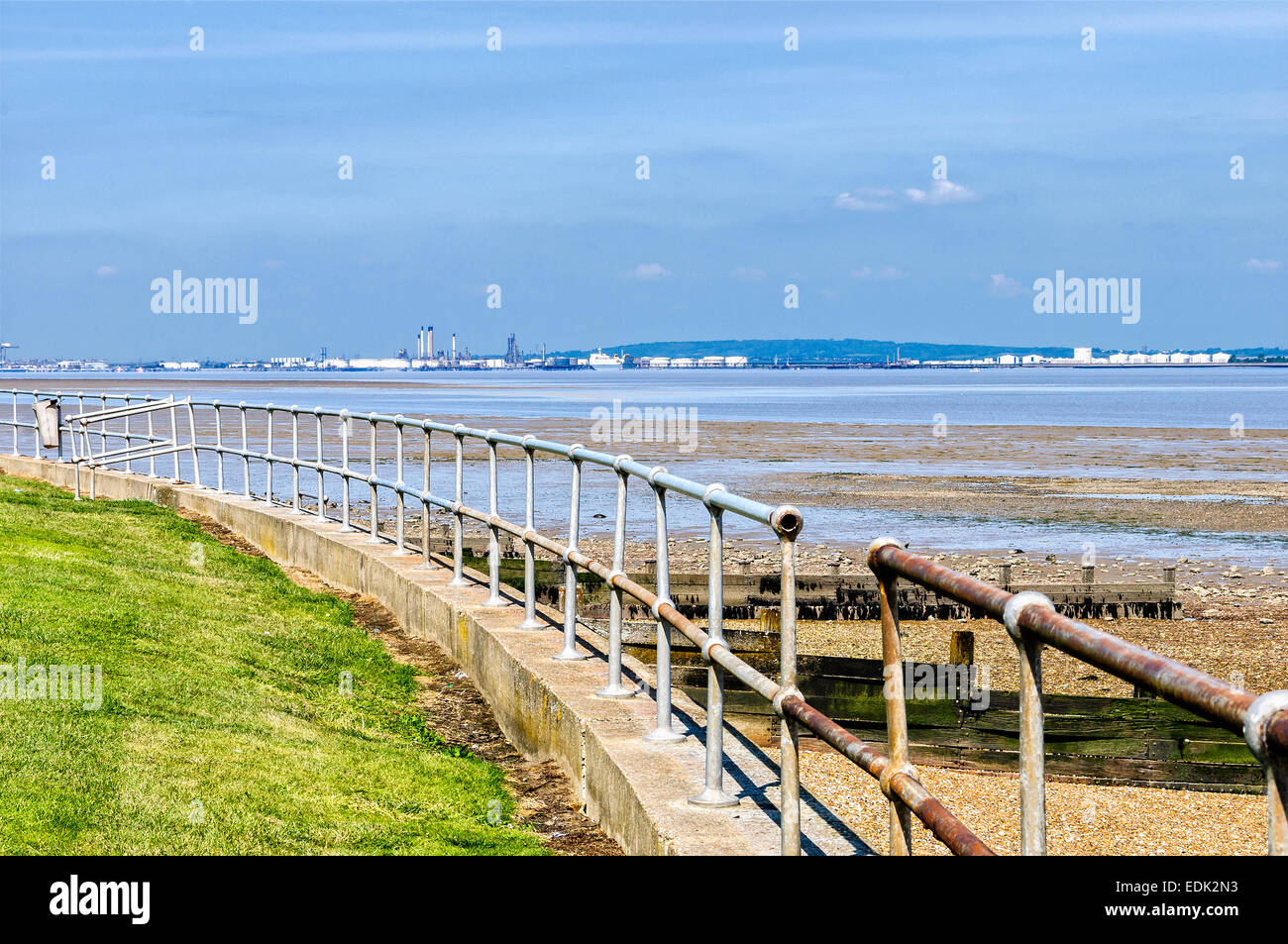 A stout rail stands on top of a sea wall to prevent falls as the sun reflects from petrochemical plant on the opposite shoreline Stock Photo