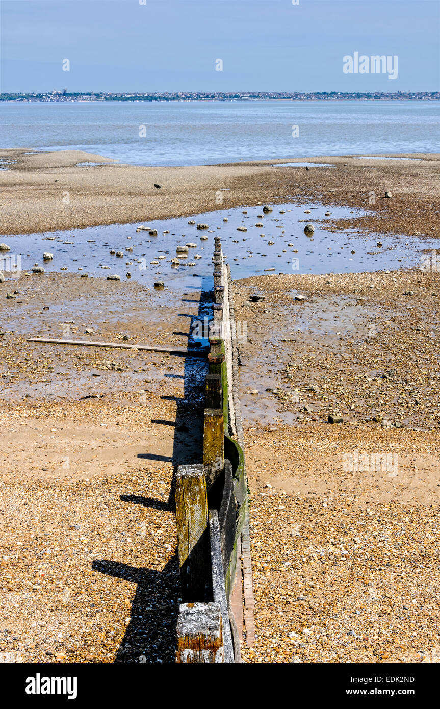 A sunlit wooden breakwater points over a rockpool towards a tranquil River Thames as it flows seawards through the estuary Stock Photo
