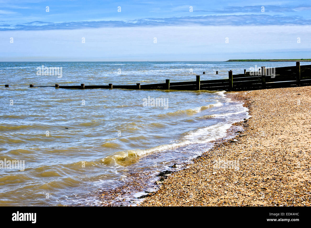 A tranquil River Thames contained by dark wooden breakwaters laps gently onto a shingle beach under a two-tone blue sky Stock Photo