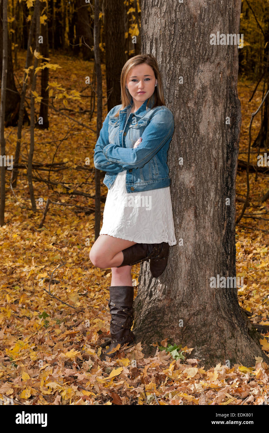 full length portrait of young female teen leaning against tree arms crossed in forest during autumn Stock Photo