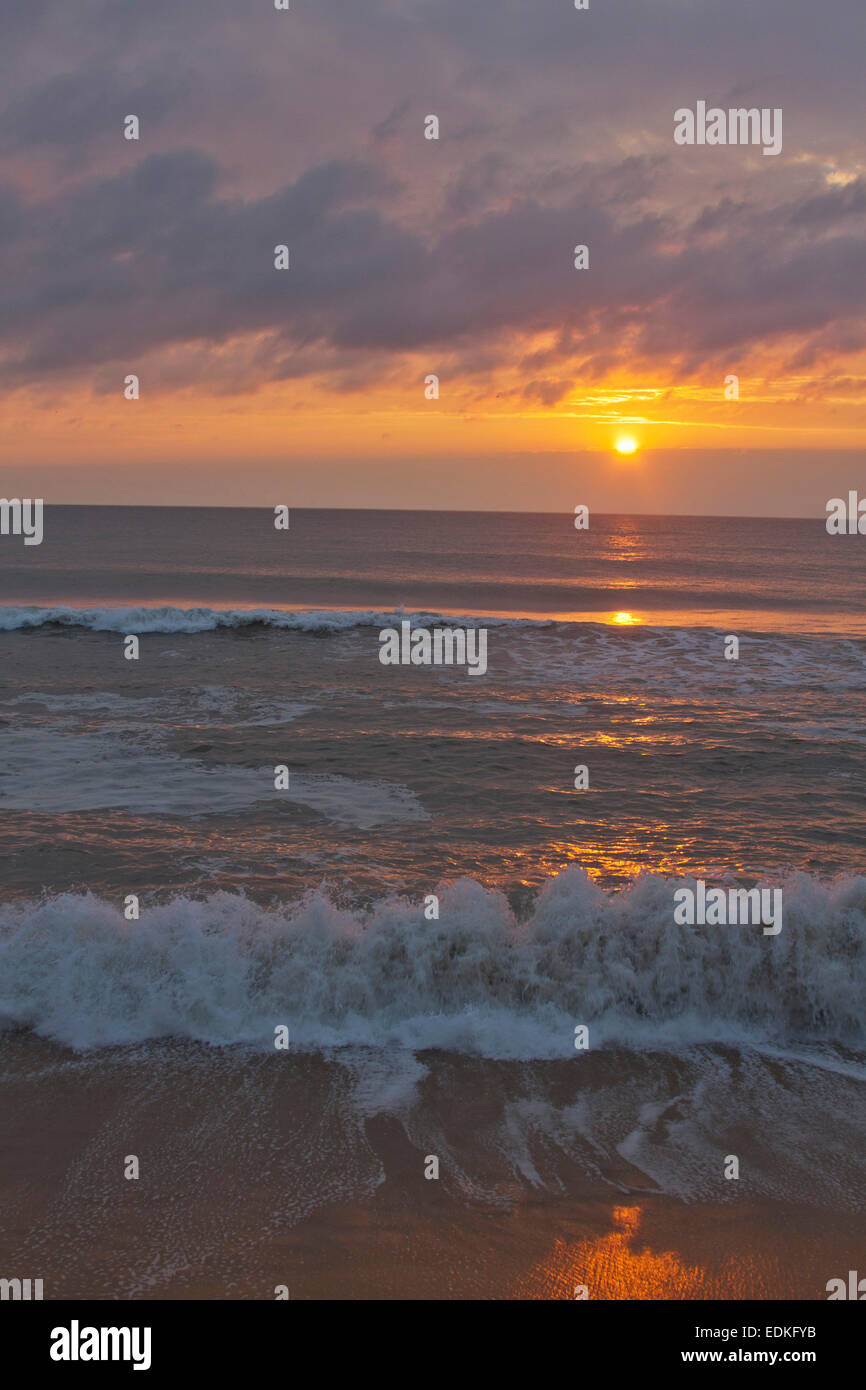 A dark, restless ocean reflects the colors of a sinking sun on a beach at twilight Stock Photo