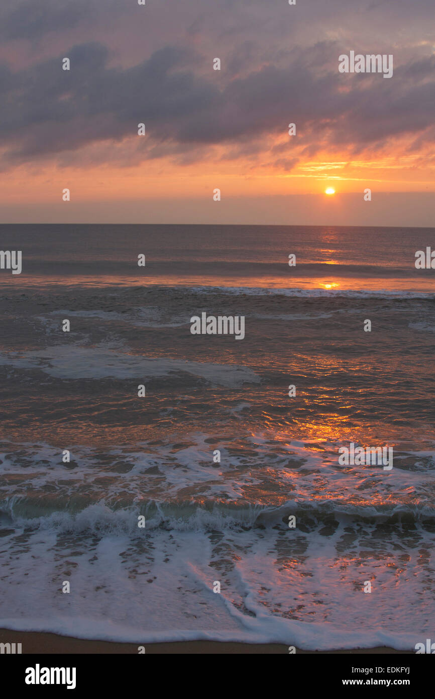 A dark, restless ocean reflects the colors of a sinking sun on a beach at twilight Stock Photo