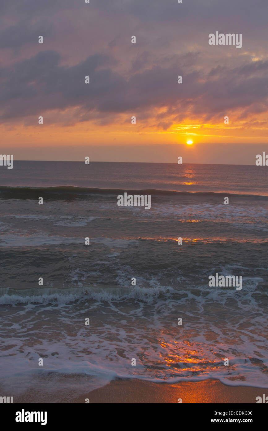 A dark, restless ocean reflects the colors of a sinking sun on a beach at twilight Stock Photo