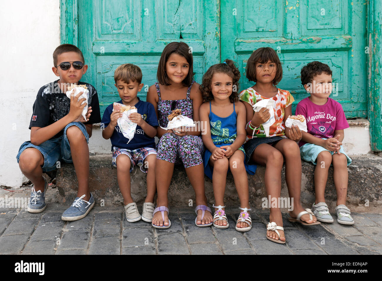 Children sitting in front of a green door holding food, Teguise, Lanzarote, Canary Islands, Spain Stock Photo