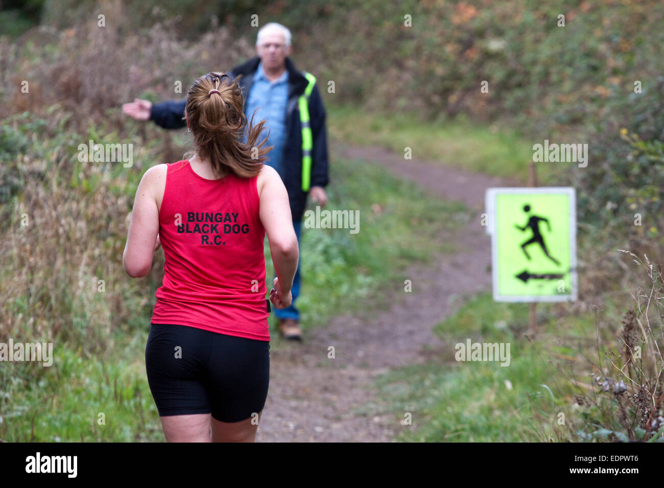 Woman runner in red top approaches a marshaled turn in a cross country race. Stock Photo