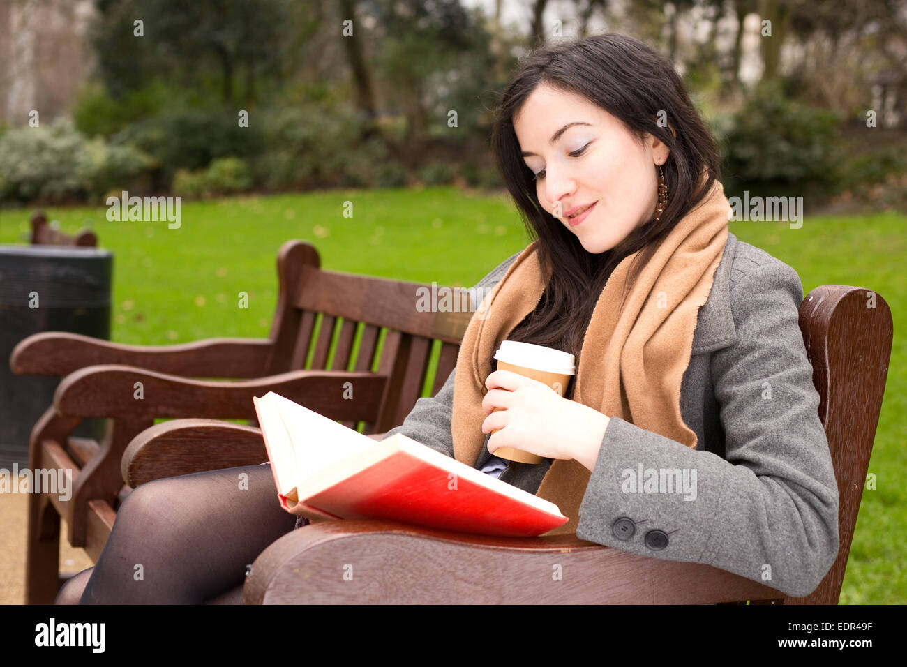 young woman sitting in the park reading a book and drinking a coffee ...