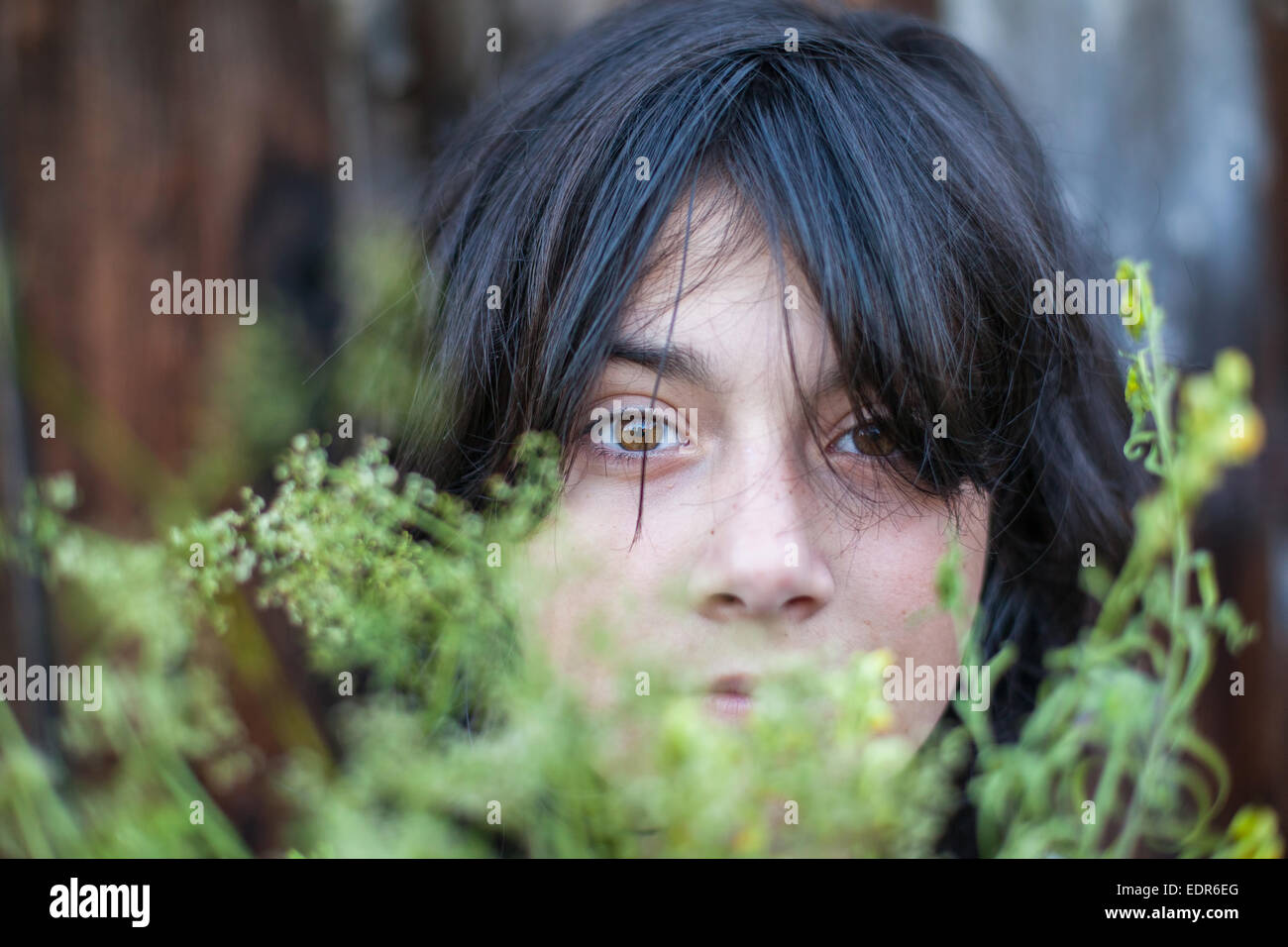 Closeup portrait of black-haired teen girl, hidden in the greenery of the garden. Stock Photo