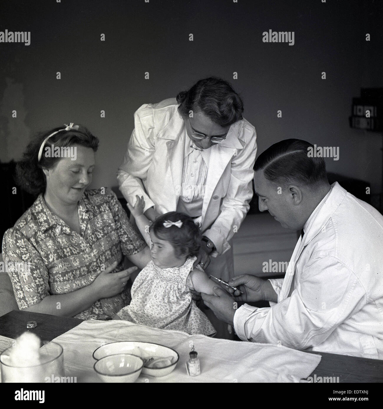 1950s, historical picture, at a GP's surgery, a doctor giving a brave young girl an injection into her left arm, the child's body is held by a nurse and with her mother is by her side in support. Stock Photo