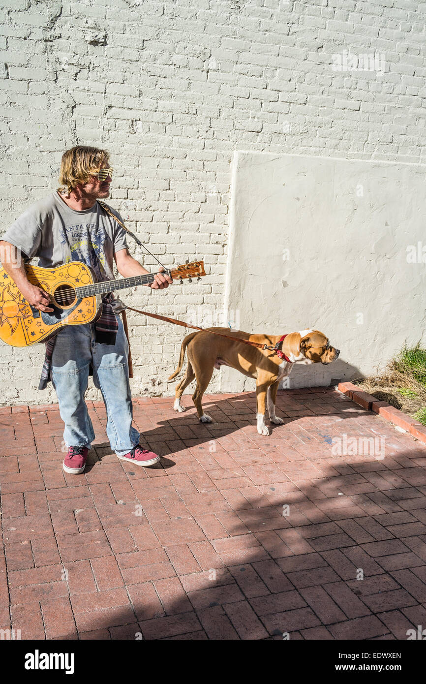 A guitar carrying man and his dog both wear sun glasses as they walk down State Street in Santa Barbara, California. Stock Photo