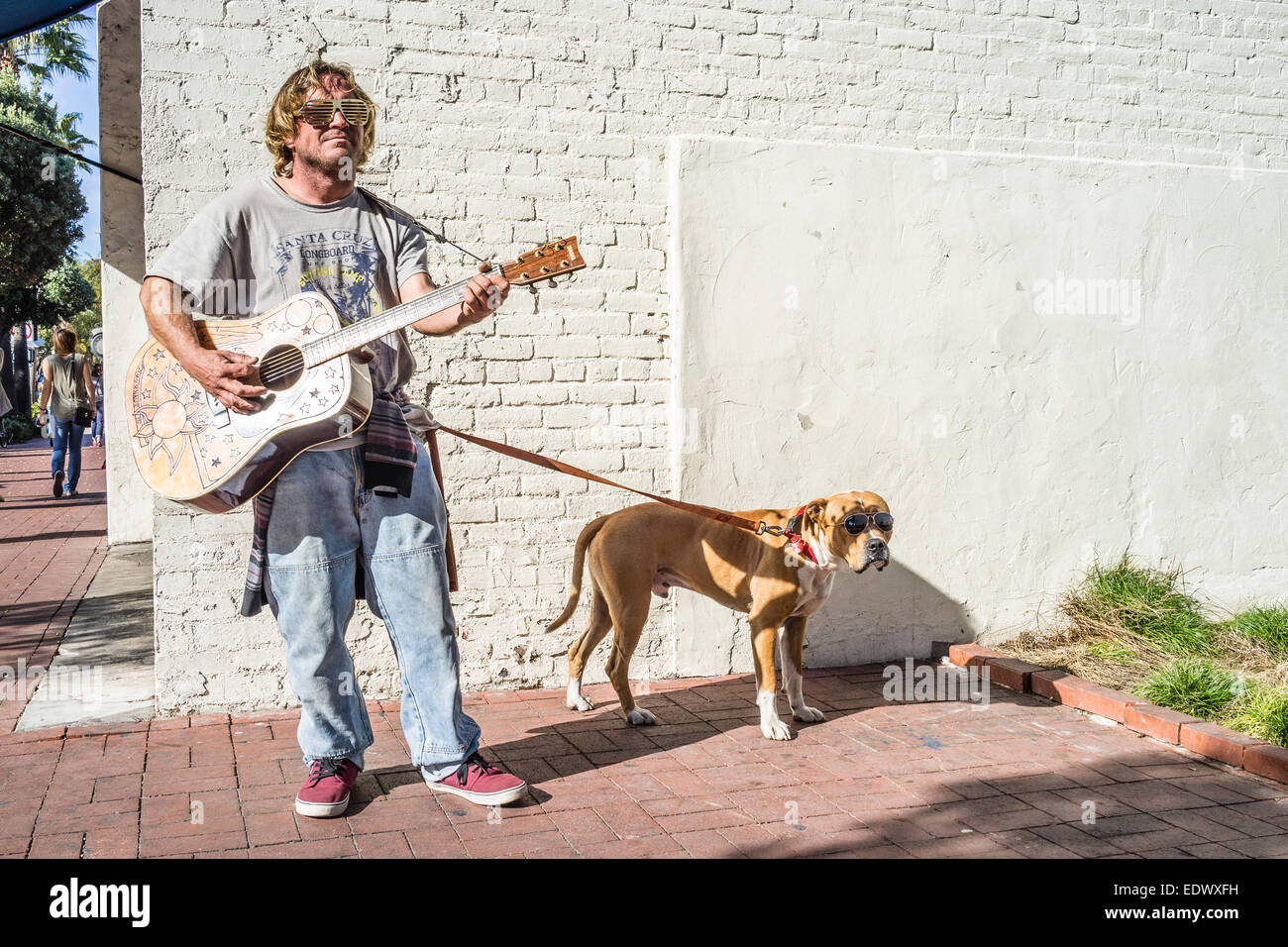 A guitar carrying man and his dog both wear sun glasses as they walk down State Street in Santa Barbara, California. Stock Photo